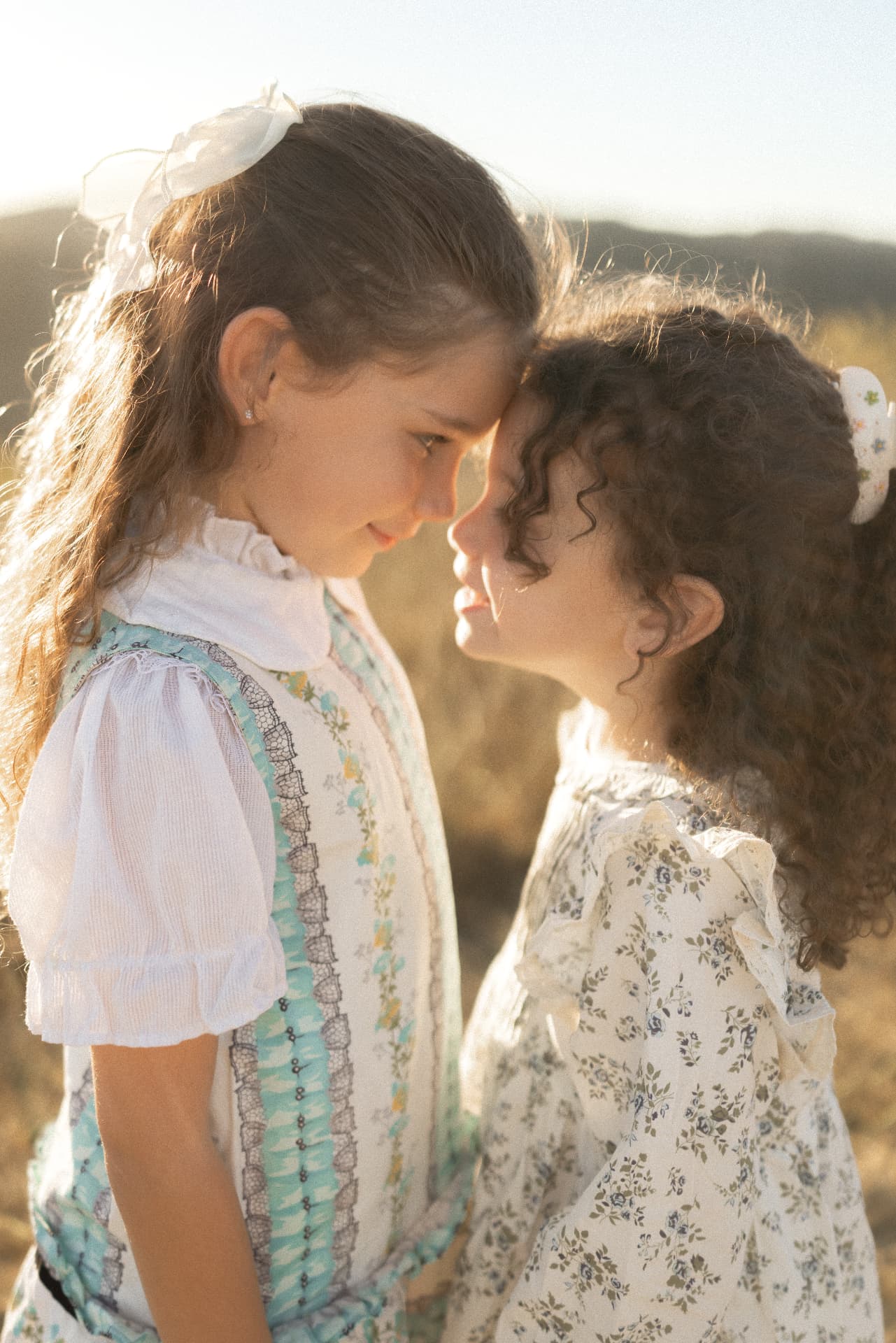Two sisters in floral dresses touching foreheads in golden light